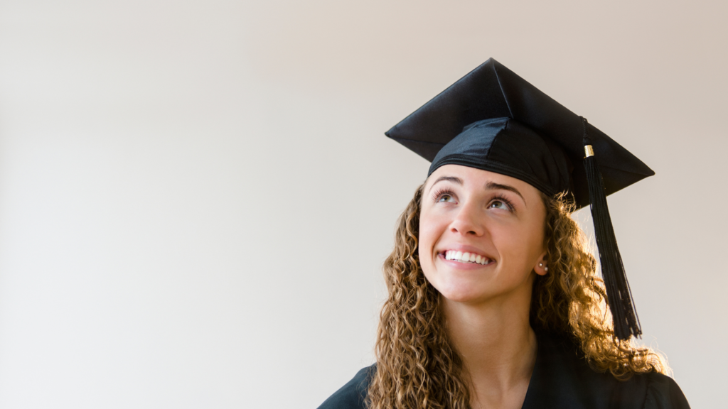 Young white woman with curly hair wearing graduation cap looking up off to one side into the distance and smiling.