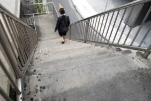 Woman with long dark hair walks down a flight of concrete steps of an overpass, no one else in sight. Viewer is looking down the steps at her near the bottom.