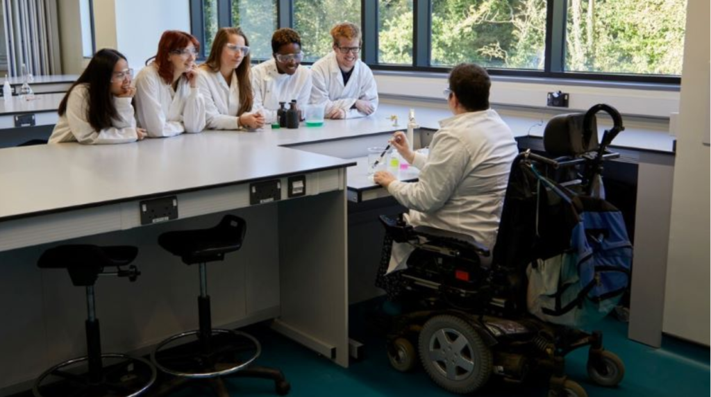 A woman with short brown hair and safety glasses sits in a large electric wheelchair at a workbench which is adjustable down to her level. She holds a pipette in a glass beaker as she looks at five students of different races wearing white labcoats and safety goggles.