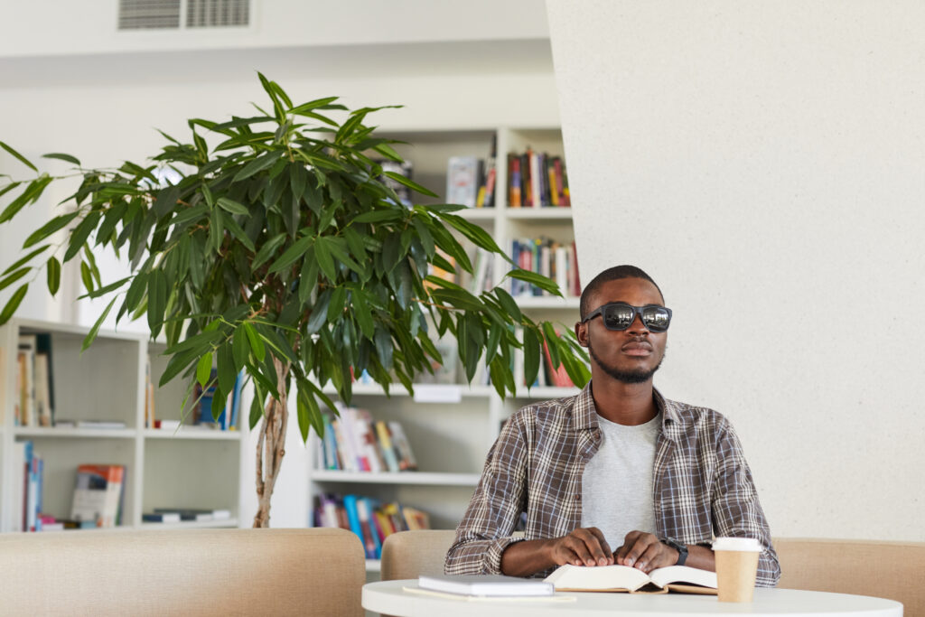 A youngish black man wearing dark glasses sits in a bright library with a small tree just behind him. He sits at a desk, looking straight ahead as both hands rest on a book, presumably Braille.