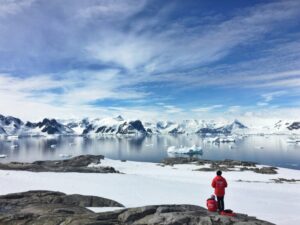 Photo of two people in red coats and woolly hats sit and stand on rock looking at a snow covered landscape and a bay of water with floating ice. The sky is bright blue with whispy clouds and snow covered mountains are in the distance.