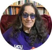 White person with long brown wavy hair past their shoulders, wearing blue-tinted glasses and a UCU t-shirt.