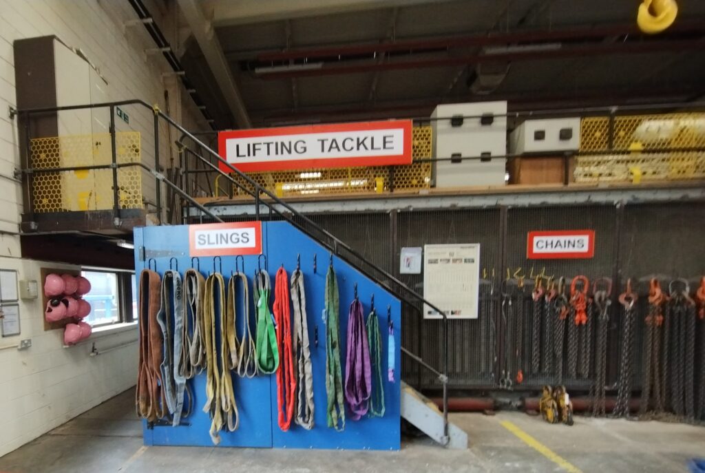 An industrial workshop has a row of hooks on which are slings, a row of hooks containing different size chains, and a big sign saying Lifting Tackle. Stairs go to an upper mezzanine level.