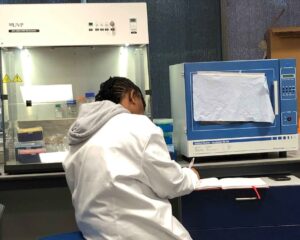 Black woman with cornrows perches on a stool writing notes in a microbiology lab