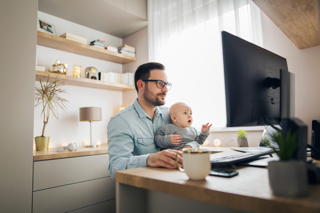 A white man with close beard and glasses sits at a home office at a computer, holding a mouse in right hand and the left hand grips a baby on his lap. The baby is looking at the computer screen in utter wonder.