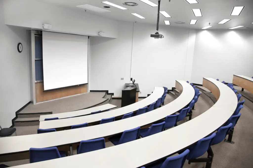 An empty lecture hall, with curved fixed desks and chairs pointing towards the corner where there's a large screen and a lecturn