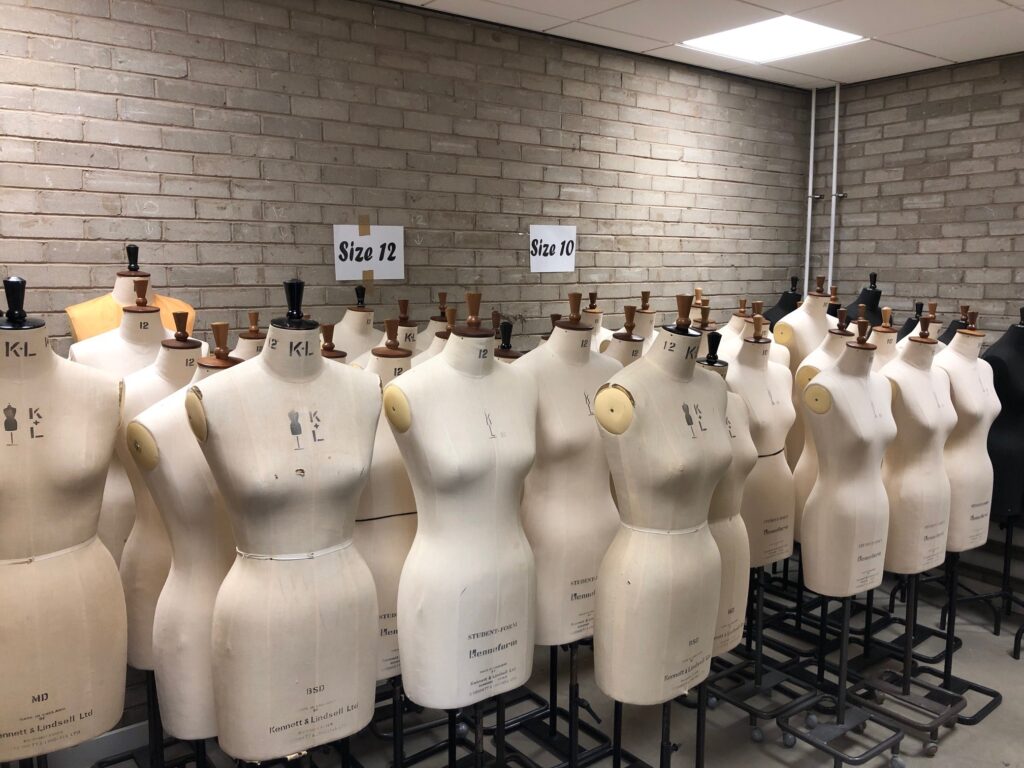 A room full of manequins, mostly of the female body, of varying dress sizes. All headless and standing on wire frames in a grey brick room.