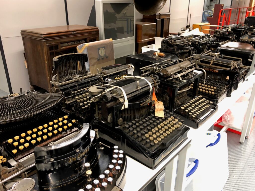 A long workbench with two rows of typewriters from the 19th and 20th centuries. They appear black with either white or faded yellow keys. Some have tags showing tied to them.