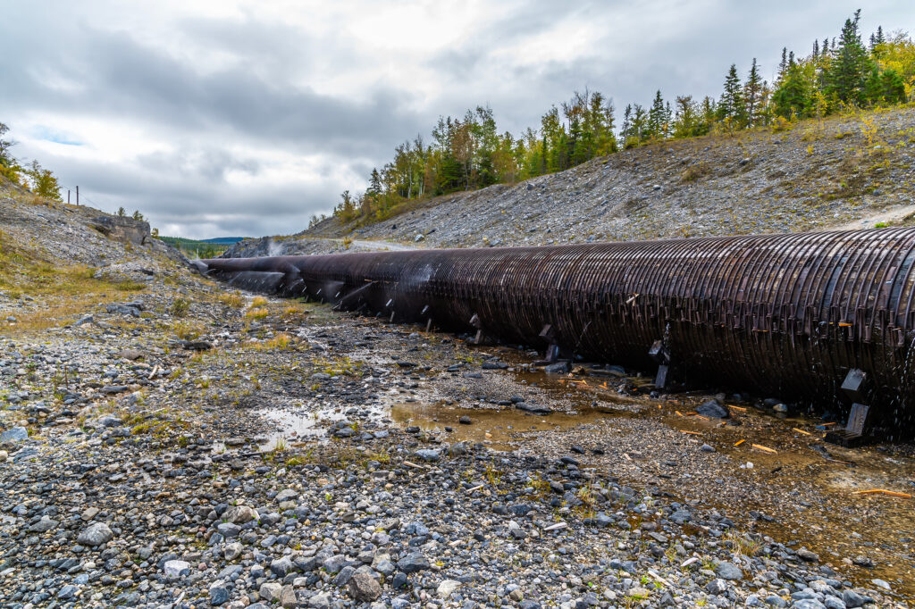 A large, rather rusty and quite leaky pipeline crosses the Canadian countryside on a gravel bed. Stubby pine trees are visible up the embankment, and puddles of water lay around the leaking pipeline.