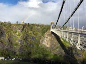 Photo of the Clifton suspension bridge, a Victorian structure high over the River Avon. Fluffy white clouds dominant the blue sky and a rainbow is visible stretching into one cloud. A greenery-covered cliff stretches down to the river, with a road running along the riverbank.