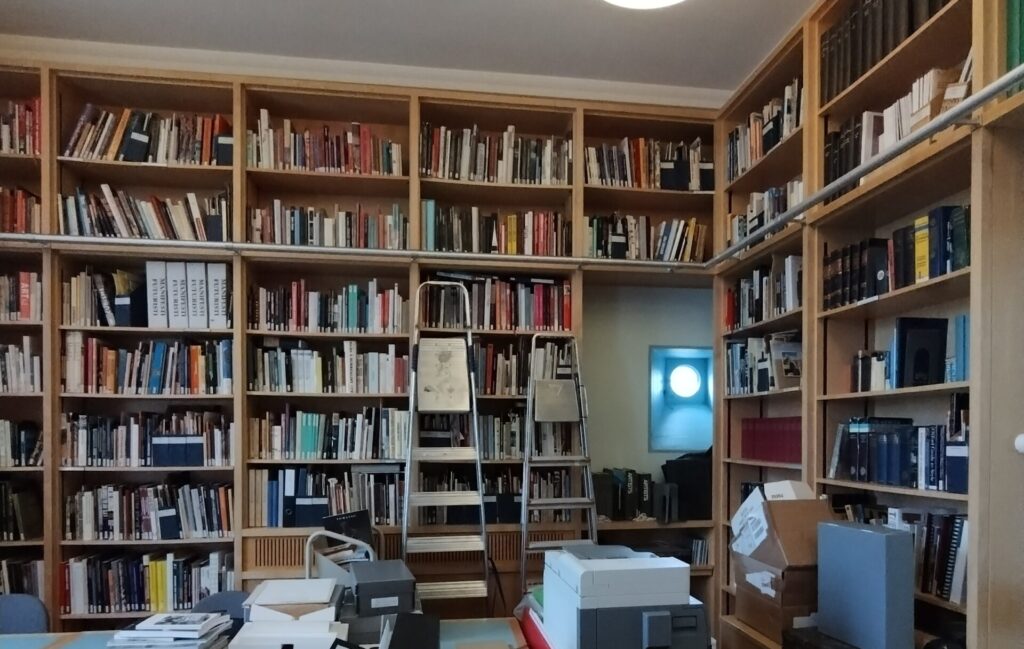 Two walls of floor to ceiling bookshelves full of books. Boxes and papers and files in on the table.