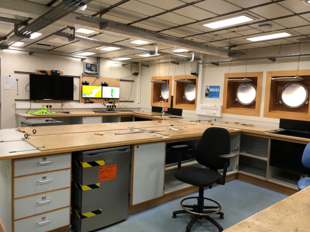 A lab on the ship with round portholes on one far wall. Rustic wooden counter tops enable equipment to be fixed to them. Under the counters are drawers, a fridge, and shelves. A black swivel chair with no wheels sits in the U shape of lab benches.