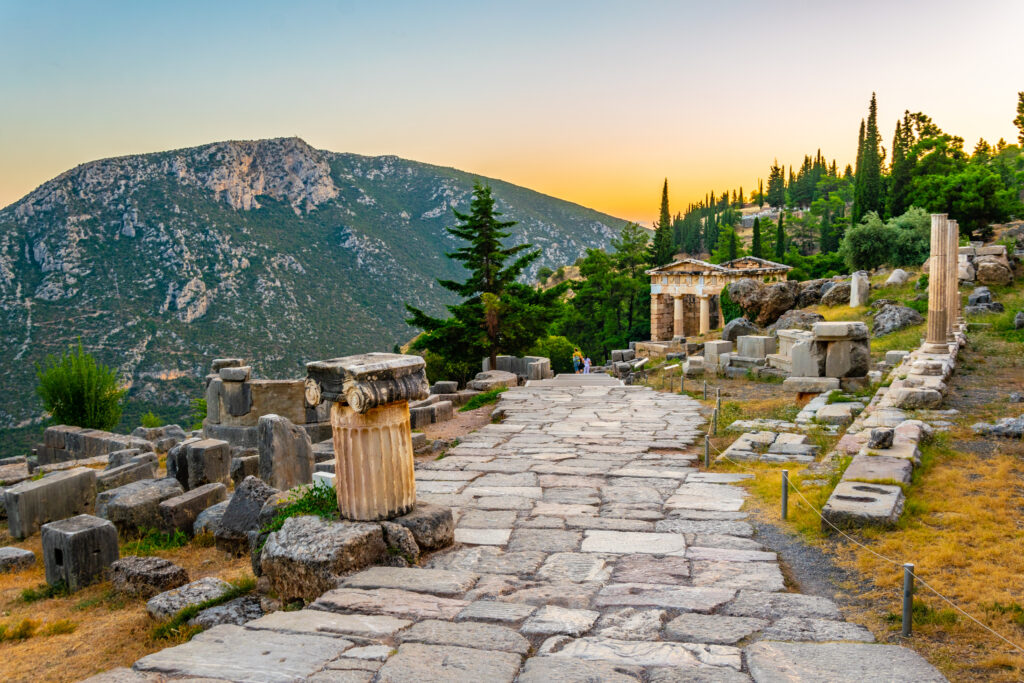 Photo of the ancient ruins of Delphi in Greece at sunset. A stone path windws down the hillside, with ruins of templs and columns on either side. A mountain looms up in the far distance across a valley. Just visible as the path dips out of sight are an adult and child holding hands walking away.