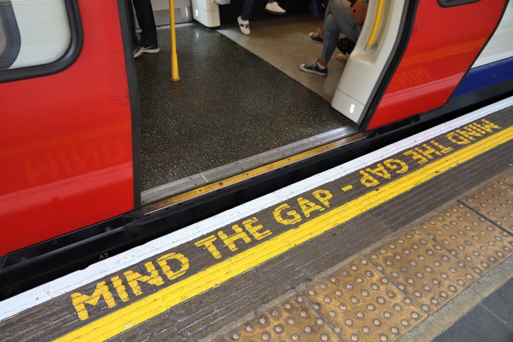 A stock image showing the London underground platform with train and open doors. Yellow lettering painted on the platform edge with iconic words Mind the Gap.