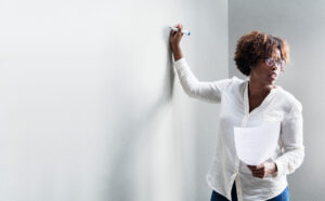 Black woman in white blouse stands looking out to the right, hand resting with pen on a whiteboard ready to write. She holds a sheet of paper in her other hand.